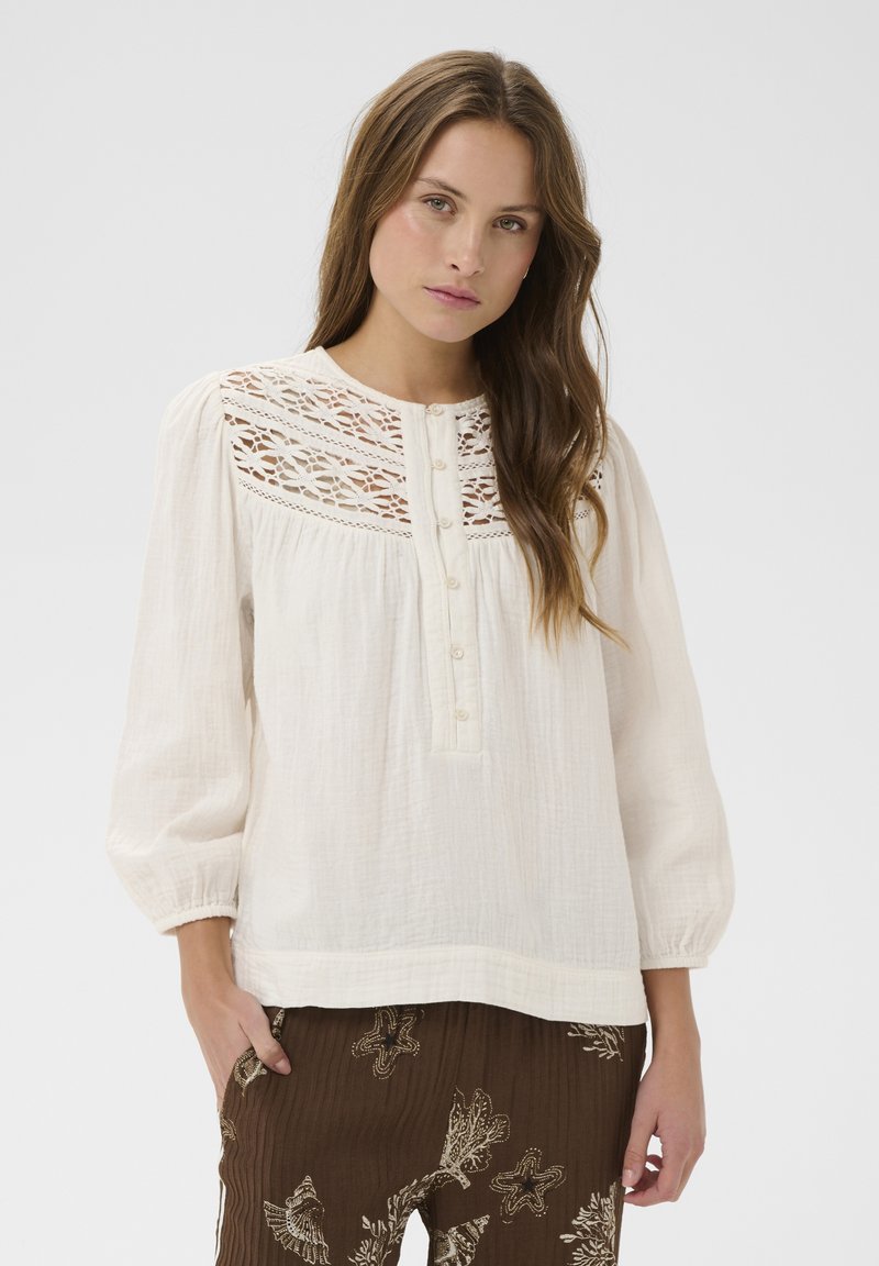 Femme aux longs cheveux bruns portant un chemisier blanc avec un empiècement en dentelle et un pantalon marron orné de motifs d'étoiles de mer et de corail, posant devant un fond uni.