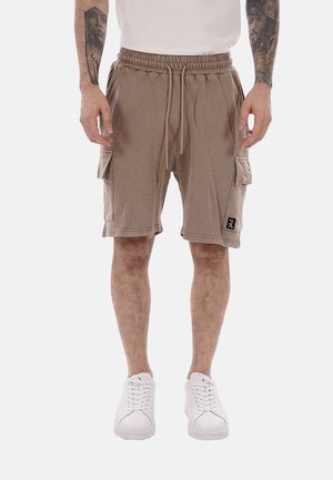 Uomo che indossa pantaloncini cargo beige con vita a coulisse, sneakers bianche e tatuaggi sulle braccia, in piedi contro uno sfondo bianco semplice.