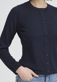 Cardigan en bleu marine côtelé avec encolure ronde, fermeture boutonnée sur le devant et manches longues. Le tissu est doux avec une texture subtile.