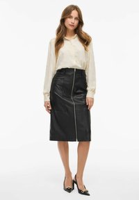 Gonna a matita nera in pelle con cerniera frontale, indossata con una blusa cream a trama. Scarpe nere con tacco e dettagli decorativi.