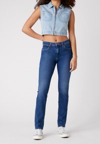 Žena oblečená ve svetle modré bezrukávové džískové sako, úzkých modrých džínách a bílých teniskách, stojící na jednobarevném pozadí.