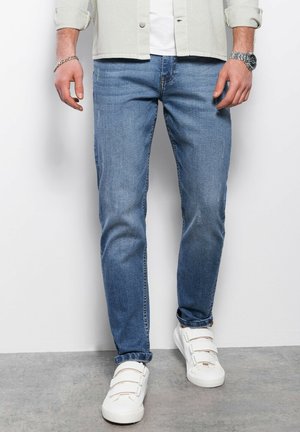 Jeans de mezclilla azul claro con corte recto, que presentan un ligero desgaste. Combinados con zapatillas blancas sin cordones y una camisa de botones gris clara.