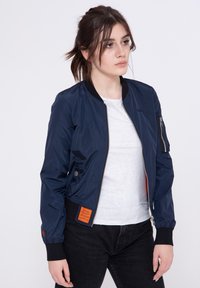 Giacca bomber blu navy in materiale lucido con cerniera frontale, polsini a coste neri e un'etichetta arancione sul davanti in basso. Maglietta grigia sotto.