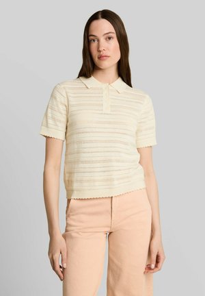 Junge Frau mit langen braunen Haaren, die ein cremefarbenes kurzes Strick-Poloshirt und hell pfirsichfarbene Hose trägt und vor einem schlichten grauen Hintergrund steht.