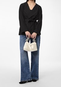 Blouse noire portefeuille à manches longues avec une ceinture, associée à un jean évasé bleu et un sac à main crème avec des anses perlées.