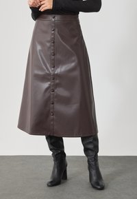 Falda midi de cuero sintético marrón con cierre de botones en la parte delantera y un acabado suave y brillante. Llevada con botas de tacón altas negras hasta la rodilla.