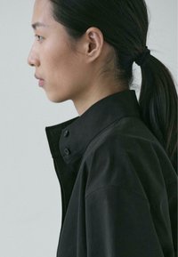 Schwarzes Hemd mit Kragen und hohem Ausschnitt, ausgestattet mit einem Knopfdetail am Kragen, hergestellt aus glattem Stoff, seitlich vor hellem Hintergrund betrachtet.