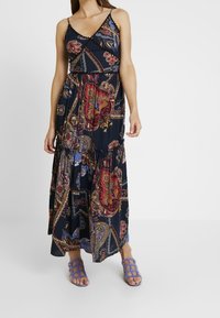 Kvinna som bär en ärmlös, ankellång klänning med mönster i flerfärgad paisley och lila sandaletter med öppen tå.