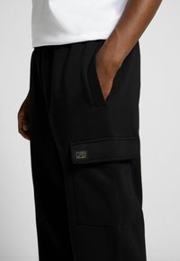 Svarta cargo-byxor med en ficka på sidan, med en liten logotagg. Tyget verkar mjukt med en slät textur och lös passform.