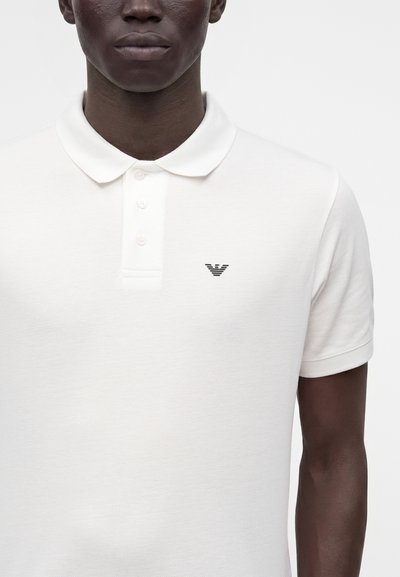 Vue de face d'une personne portant un polo blanc à manches courtes avec trois boutons et un petit logo noir sur la poitrine gauche.