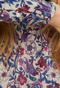 Blomstret stof har en lys beige baggrund med multicolore paisley- og blomsterdesigns i lilla, blå og pink nuancer.