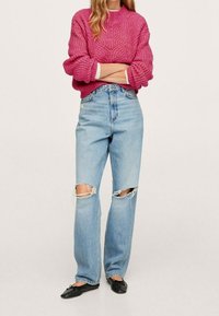 Pull en tricot rose avec un col montant, associé à un jean bleu clair déchiré avec des genoux effilochés et une coupe décontractée. Des ballerines noires complètent le look.