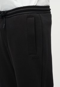 Μαύρη υφασμάτινη φόρμα με πλαϊνή τσέπη, ελαστική μέση, κορδόνι και διακριτικό λογότυπο BOSS κοντά στην τσέπη, σε άτομο που φοράει λευκό πουκάμισο.