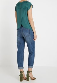 Tyrkysový top s krátkým rukávem a detailním střihem na zádech, kombinovaný s džíny z modrého denimu s vysokým pasem. Olivově zelené sandály na podpatku dokončují outfit.