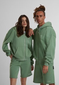 Ensemble sport en molleton vert composé d'un hoodie et d'un short, doté d'une fermeture éclair, de poches avant et d'un logo brodé. Texture douce, coupe décontractée.