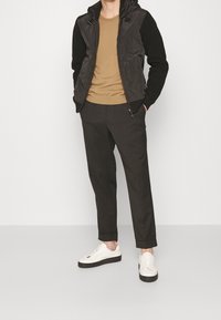 Chaqueta acolchada negra con capucha y mangas de punto oscuras, camisa marrón, pantalones oscuros y zapatillas blancas con suelas negras, de pie contra un fondo blanco.