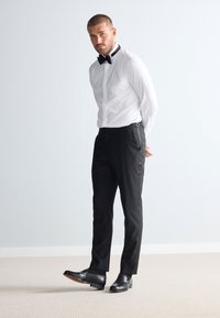 Camicia bianca, papillon nero e pantaloni neri con bretelle. Scarpe in pelle nere. Design semplice, forma aderente, texture liscia.