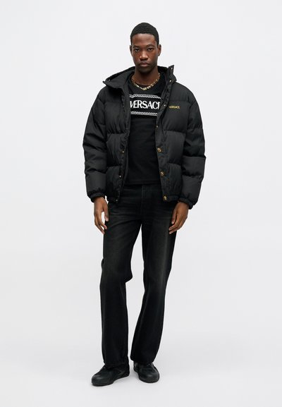 Melnas pūka jakas ar kapuci, ar spīdīgu tekstūru un zelta logo akcentiem. Valkāta virs melna T-krekla ar baltu grafisko dizainu. Melni džinsi.