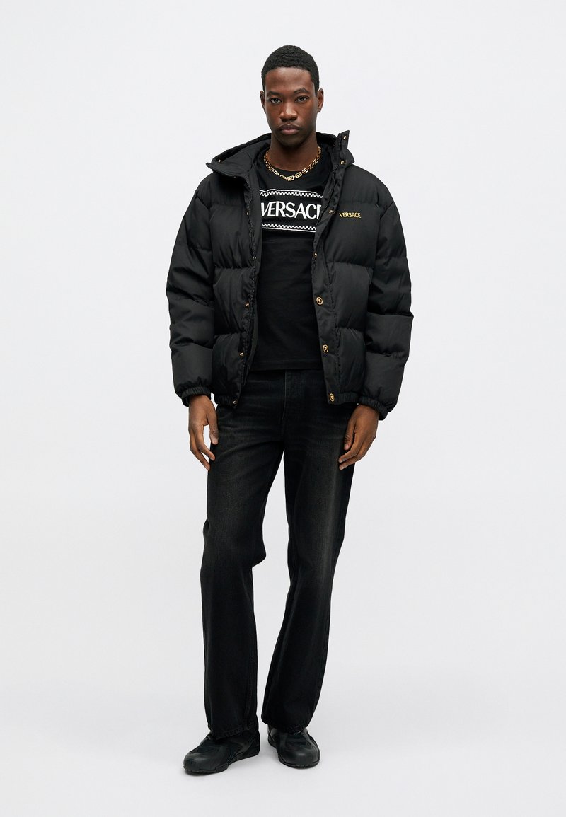 Sort pufferjakke med hætte, der har en blank overflade og gyldent logoaccent. Bæres over en sort t-shirt med hvid grafisk design. Sorte jeans.