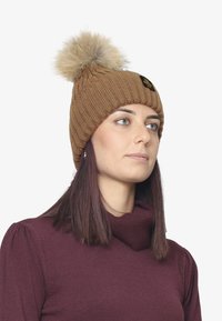 Beanie lavorata a maglia marrone con motivo a coste e un pompon in pelliccia sintetica beige sopra. Presenta un piccolo logo nero sul davanti.