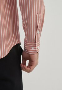 Chemise à manches longues en corail avec des rayures verticales blanches. Équipée de poignets à boutons et d'un ourlet arrondi. Tissu texturé visible.