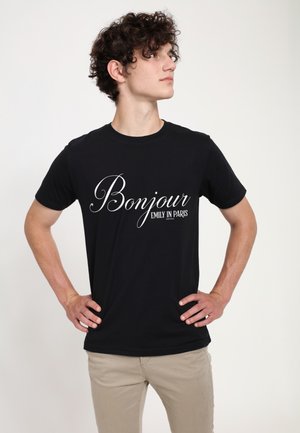Giovane uomo con capelli ricci in piedi con le mani sui fianchi, indossa una T-shirt nera con la scritta "Bonjour Emily in Paris" e pantaloni beige su sfondo bianco.