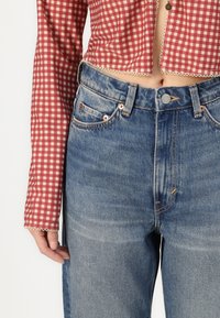 Röd och vit rutmönstrad crop top med långa ärmar; matchad med högmidjade blå denimjeans med kopparnitar och lätt blekning.