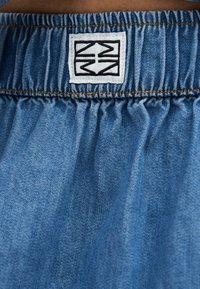 Κοντινό πλάνο μπλε τζιν σορτς με ελαστική μέση, καφέ ραφές και ένα λευκό τετράγωνο ετικέτα με μαύρα γεωμετρικά βέλη.