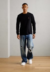 Sort langærmet skjorte, lyseblå ripped jeans og hvide sneakers. Skjorten har et lille emblem, og jeansene er løstsiddende.