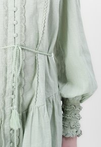Hellgrünes, besticktes Kleid aus strukturiertem Stoff, mit Knopfleiste, gerafften Ärmeln und gebundenem Taillengürtel. Verfügt über dekorative Akzente und Rüschen.