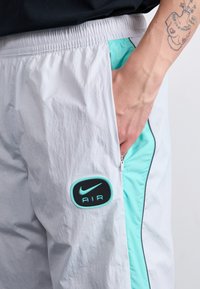 Nike Air kalhoty ve světle šedé barvě s kontrastním aqua bočním panelem, obsahující kapsu na zip a nášivku s logem na stehně.