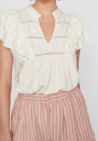 Blusa beige chiara con maniche a volant, scollatura a V e dettagli in pizzo. Abbinata a pantaloni a strisce rosa, plissettati, con vita elastica.