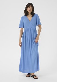 Maxikleid aus hellblauem Stoff mit strukturiertem Muster, V-Ausschnitt, kurzen Ärmeln, gerafftem Bund und fließendem Rock. Kombiniert mit schwarzen Sandalen.