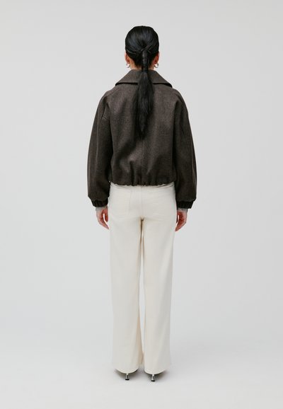 Giacca bomber in misto lana marrone con polsini a costine, abbinata a pantaloni ampi color crema e accessori in argento. Lo sfondo è semplice e neutro.