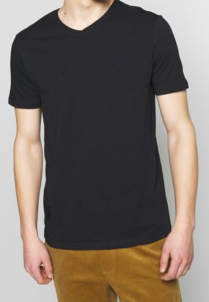 Man wearing plain black short-sleeve V-neck T-shirt and brown corduroy pants, standing against a plain gray background.