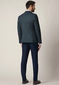 Mężczyzna w ciemnozielonym żakiecie, granatowych spodniach, brązowych eleganckich butach, stojący tyłem na tle jasnobeżowym.