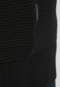 Svart stickad tröja med ribbad textur och kontrasterande vertikala ränder på sidan. Inkluderar en liten vävd etikett med detaljer.