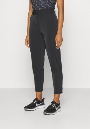 Person iført sorte taperede bukser, sorte og hvide Nike-sneakers og en mønstret sort top mod en ensfarvet baggrund.