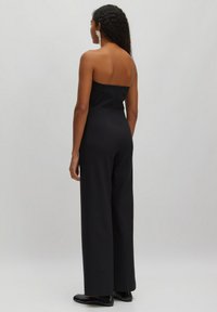 Zwart strapless jumpsuit met een fitted lijfje, wijde broekspijpen en een gladde textuur. Geaccentueerd met grote, metallic oorbellen.