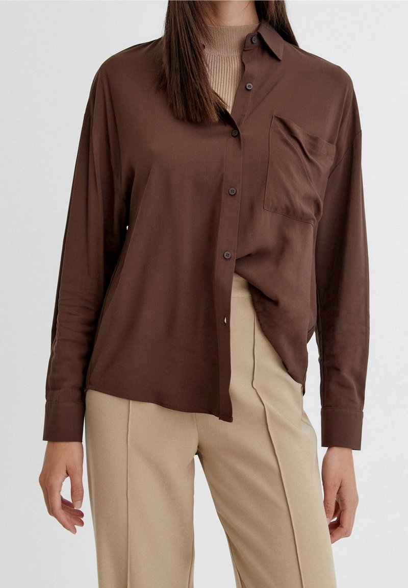 Femme portant une chemise marron à manches longues boutonnée portée sur un col roulé beige, associée à un pantalon beige sur un fond uni.