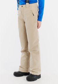 Beige sneeuwbroek met verstelbare bretels, een ritszak en een losse pasvorm. Gecombineerd met zwarte waterdichte laarzen.