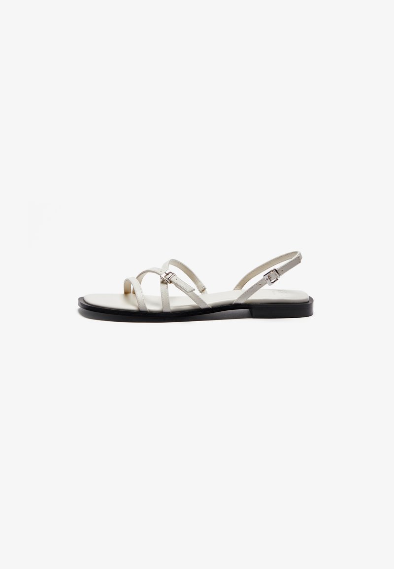 Flat sandals featuring thin metallic straps, a light cream upper, and a black sole. Adjustable buckle detail on the ankle strap.