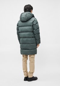 Vihreä puhvitakki hupulla, jossa on tikattu muotoilu, keskikokoinen pituus ja sileä pinta, yhdistettynä beigeihin housuihin ja mustiin saappaisiin.