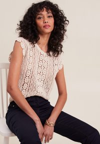 Beige gehaakte top met korte mouwen, bloemenpatroon en gegolfde randen. Gepaarde met donkerblauwe broeken en een gevlochten gouden armband.