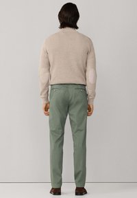 Beige gebreide trui met elleboogstukken, gecombineerd met groene broek. De broek heeft zijzakken en een rechte pijp. Bruine schoenen.