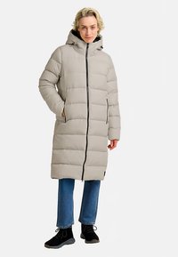 Lichtgrijze pufferjas met capuchon, voorzien van een voorrits, verticale naden en zijzakken. Draagt over een blauwe spijkerbroek en zwarte schoenen.