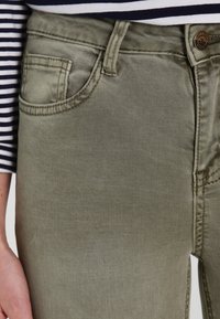 Primer plano de vaqueros de mezclilla verde oliva con un bolsillo delantero, botón, y presillas para el cinturón, combinados con una camisa a rayas azul marino y blanca.
