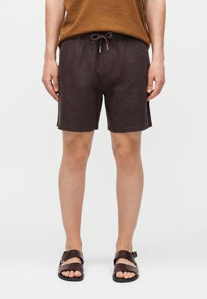 Mężczyzna w ciemnobrązowych szortach ze sznurkiem, brązowym topie bez rękawów i ciemnobrązowych skórzanych sandałach stojący na jednolitym tle.