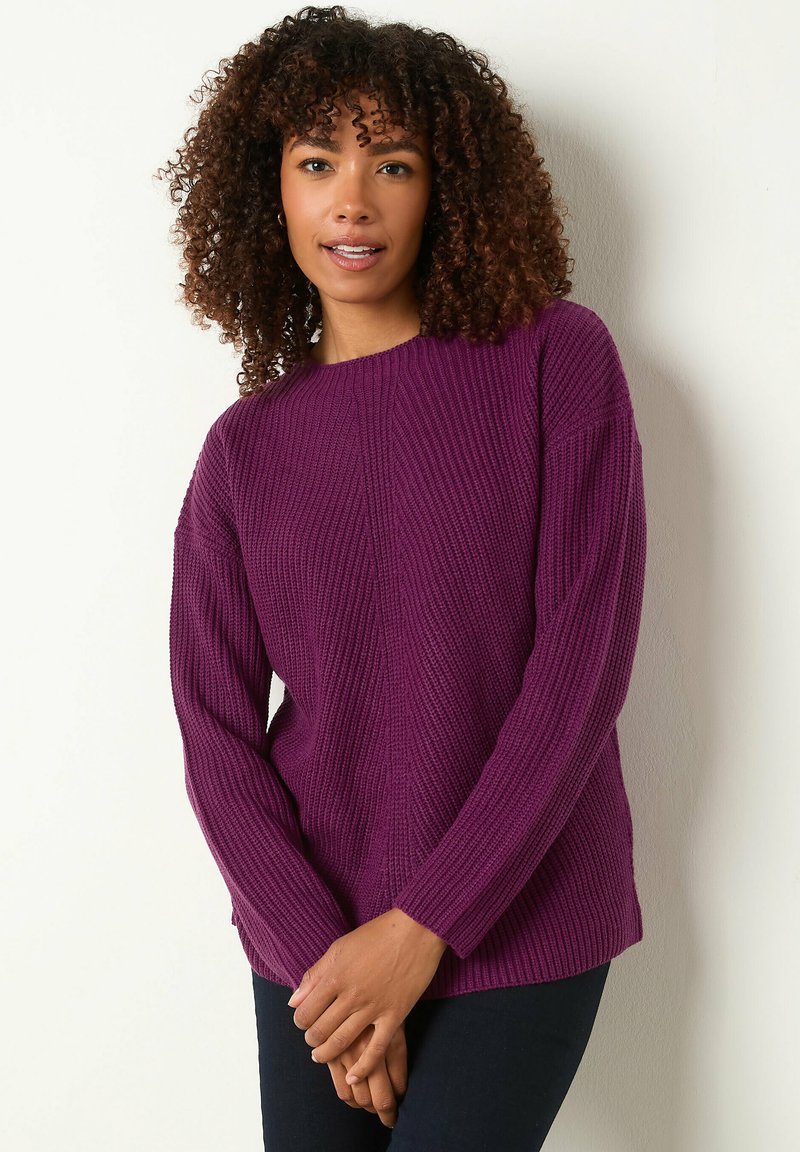 Vrouw met krullend haar, gekleed in een paarse gebreide trui en donkere broek, staande tegen een witte muur met handen voor zich samengevoegd.