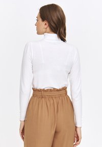 Biała golfowa sweter z długimi rękawami, zestawiony z brązowymi spodniami o wysokim stanie z falbaną w pasie.